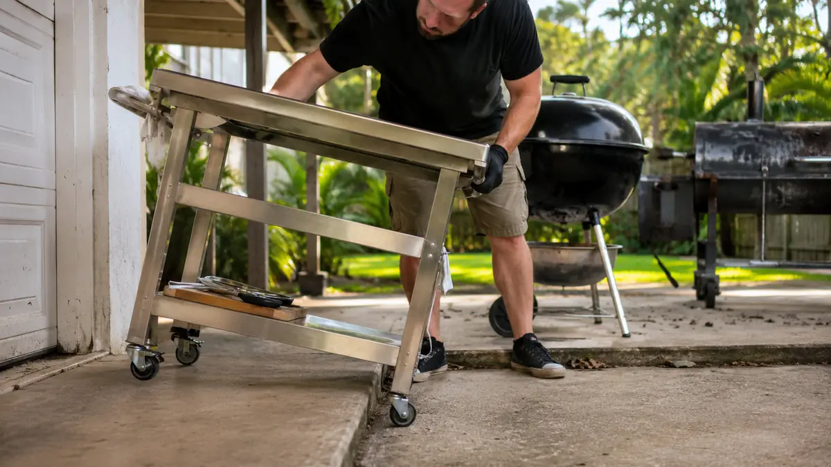 A stainless top rolling cart with its front caster catching on a raised concrete seam between a patio slab and a garage walkway