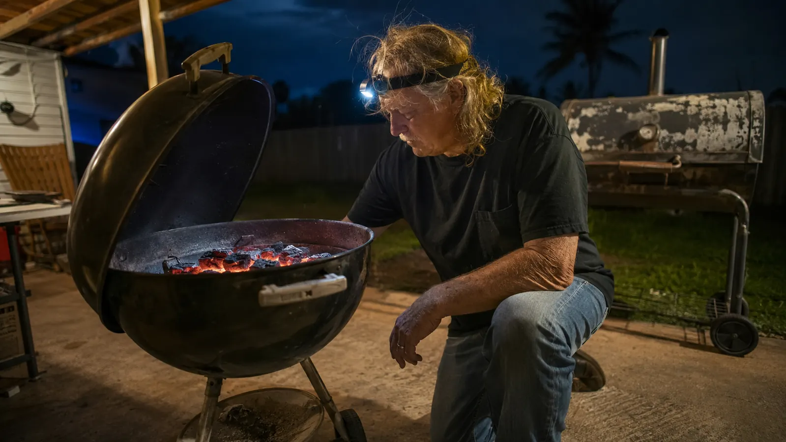 A man in a headlamp kneeling beside a charcoal kettle grill on a dark patio at two in the morning
