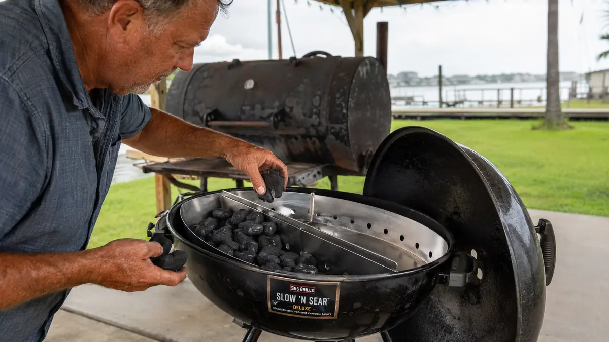 Hands loading charcoal into the stainless Slow 'N Sear basket insert inside a kettle grill